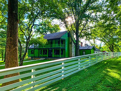 A green farmhouse behind a tree-lined white wooden fence.